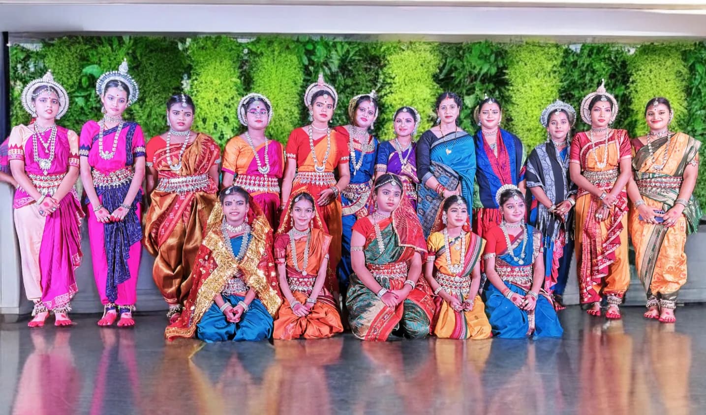 Group portrait of the academy's dancers in silver jewellery and silk costumes against a green wall