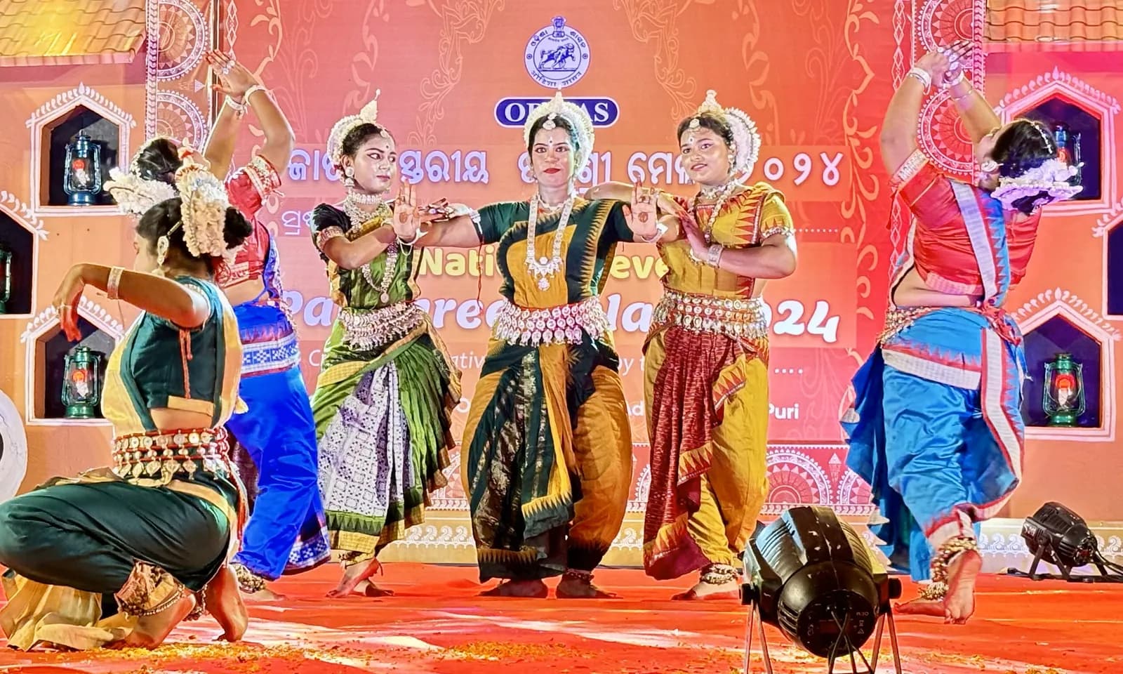 Six dancers in a tiered formation with arms raised on a festival stage